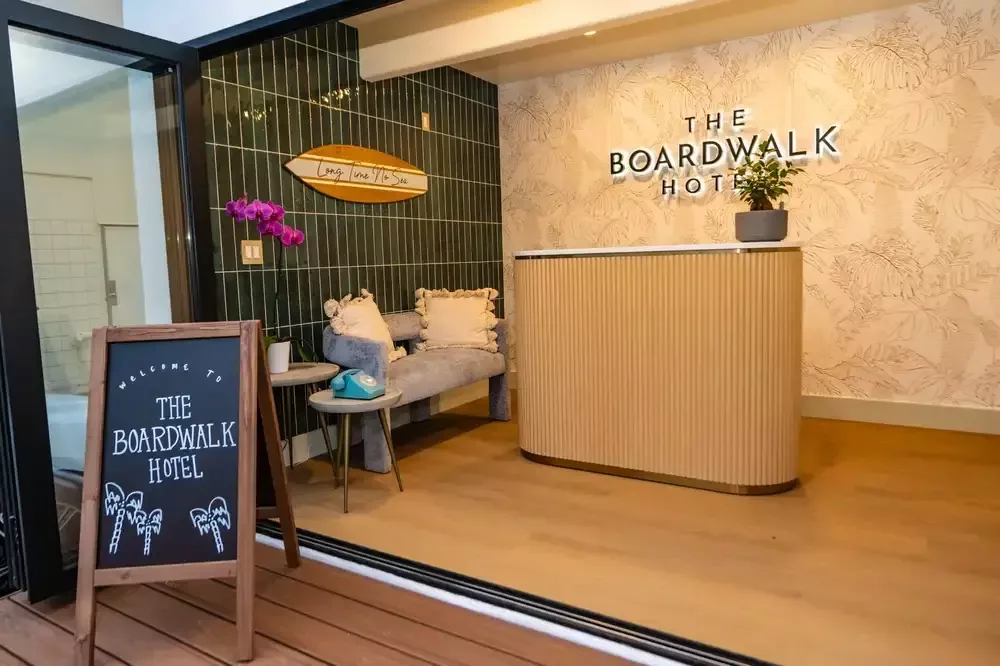 After: the Boardwalk Hotel lobby with a curved wood reception desk, wave-pattern wallpaper and a chalkboard welcome sign.
