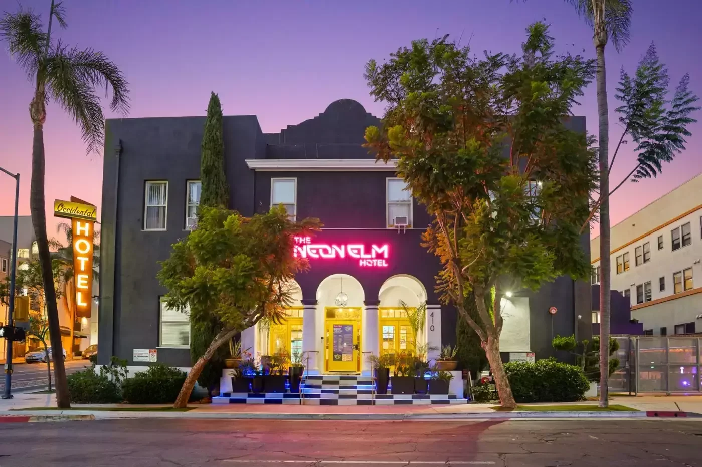 After: the Neon Elm Hotel at twilight, charcoal facade, pink neon sign, lit arches and yellow doors.