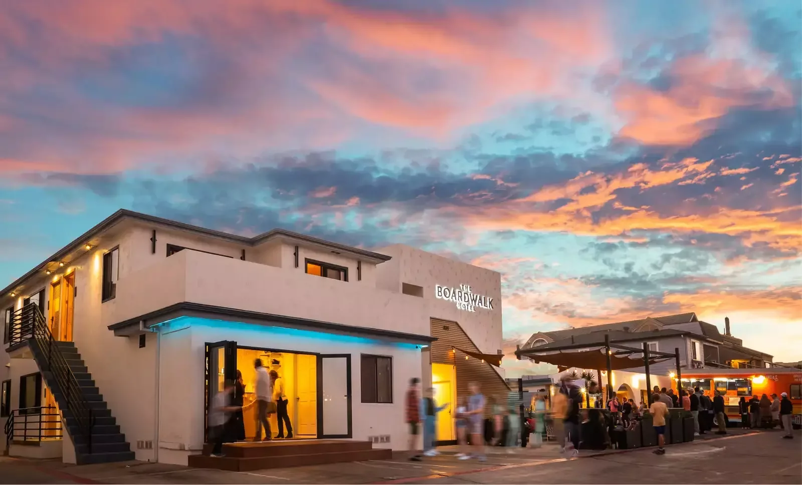 The Boardwalk Hotel in Mission Beach at sunset, guests gathered on the patio under a pink sky.